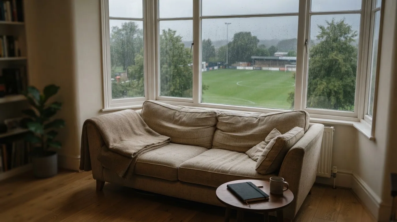 Ein Sofa mit Wolldecke steht vor einem grossen Fenster, draussen Regen und in der Ferne ein Fussballplatz.