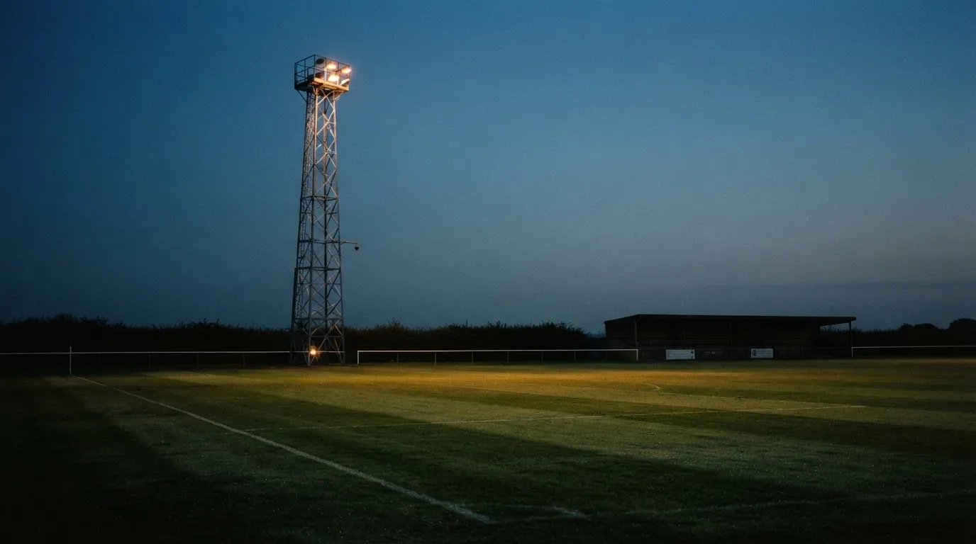 Ein Flutlichtmast ragt in den Abendhimmel, die Rasenfläche darunter liegt leer und dunkelgrün da.