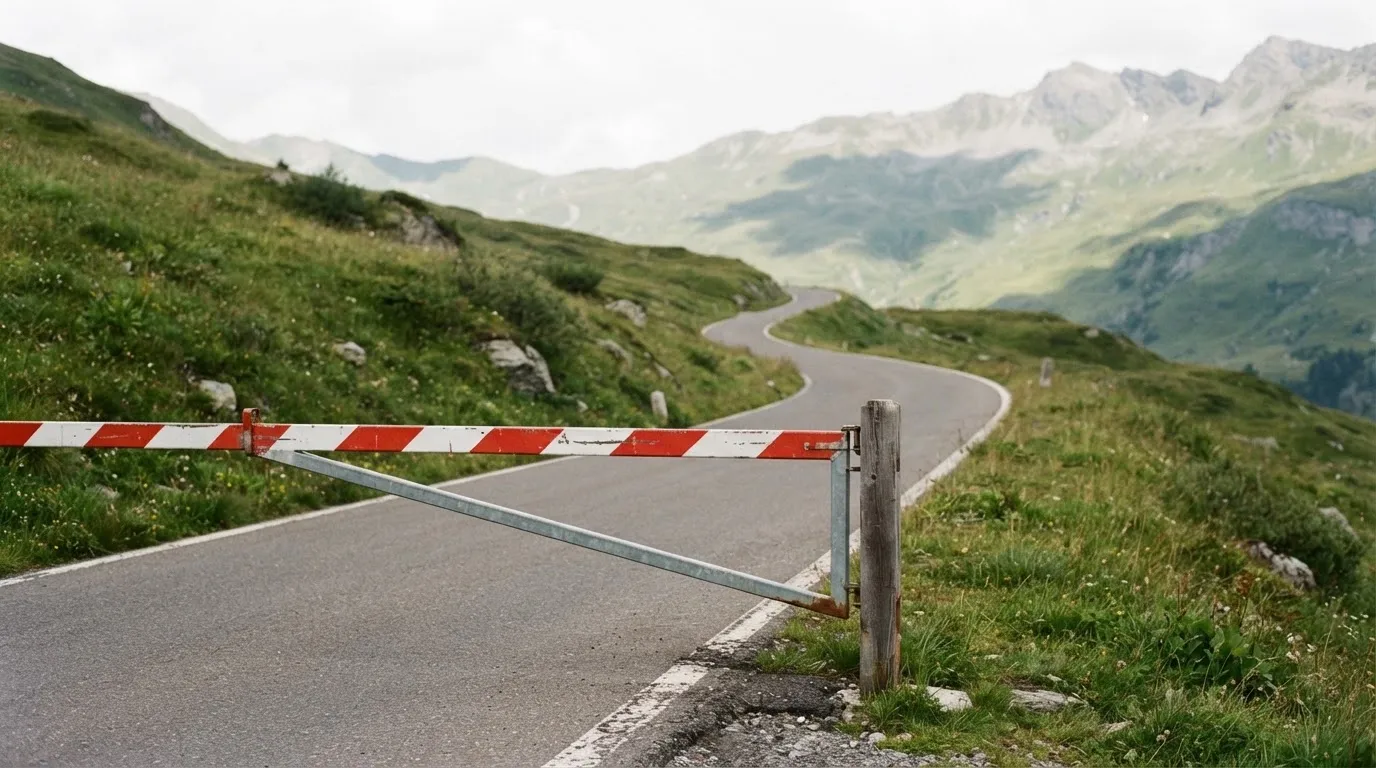 Ein Schlagbaum sperrt eine Landstrasse in den Bergen, dahinter windet sich der Weg zwischen grünen Hängen.