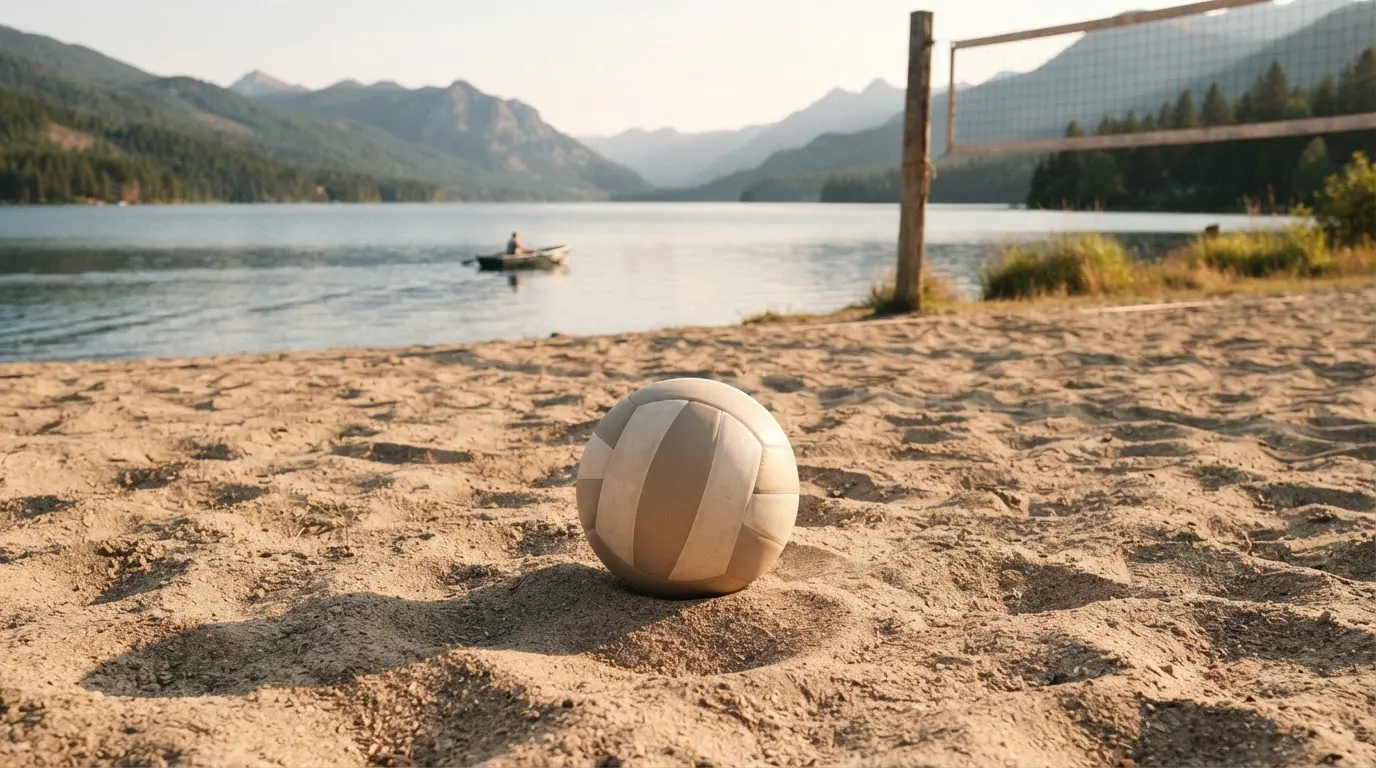 Ein Volleyball liegt im Sand eines Spielfelds am Seeufer, im Hintergrund gleitet ein Boot über das Wasser.