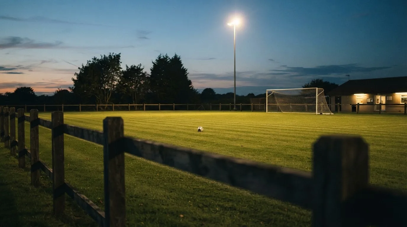 Ein kleiner Dorf-Fussballplatz liegt in der Dämmerung unter einem hohen Flutlichtmast, am Spielfeldrand steht ein Holzzaun.