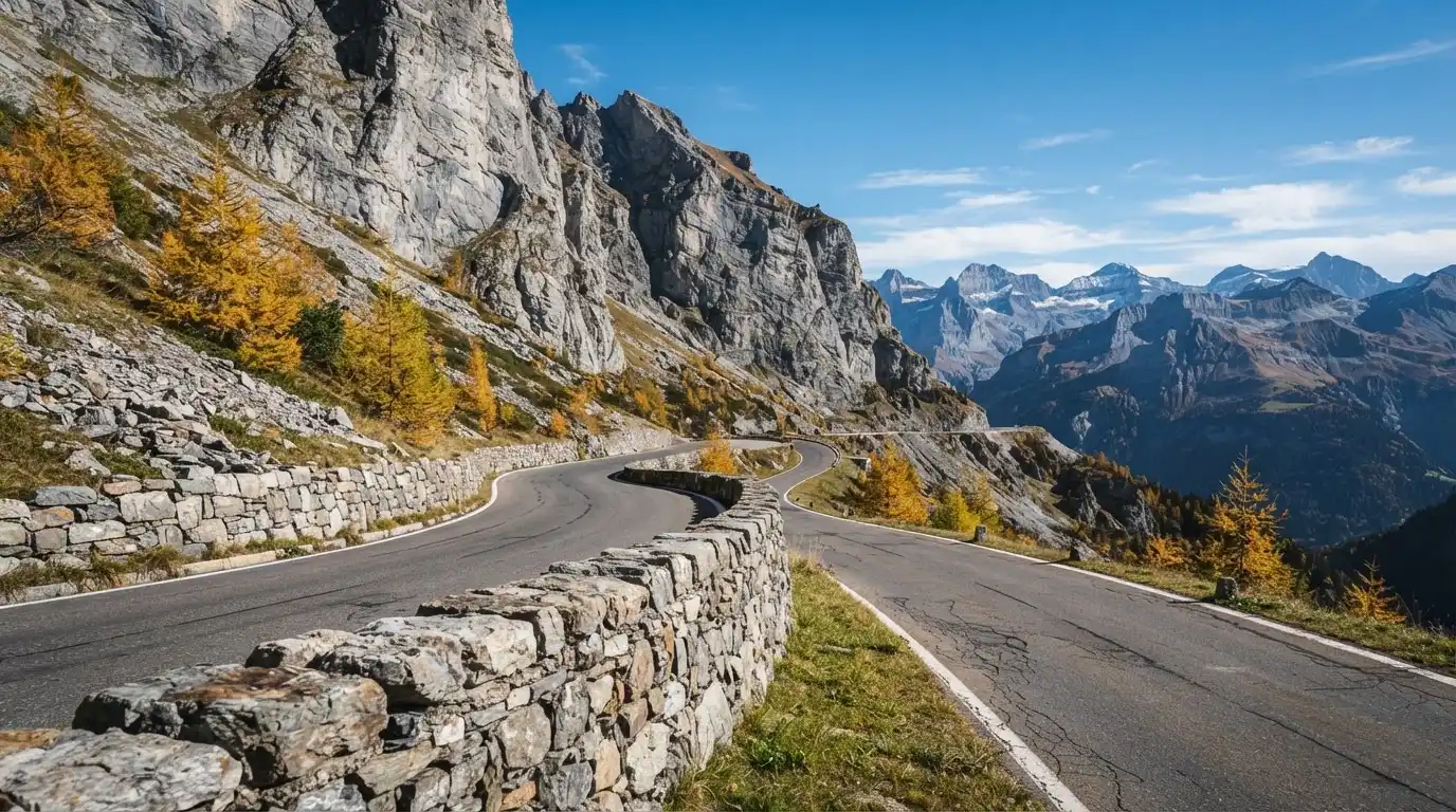 Eine Passstrasse im Hochgebirge säumt eine niedrige Steinmauer, dahinter ragen Felswände auf und der Herbsthimmel ist klar.