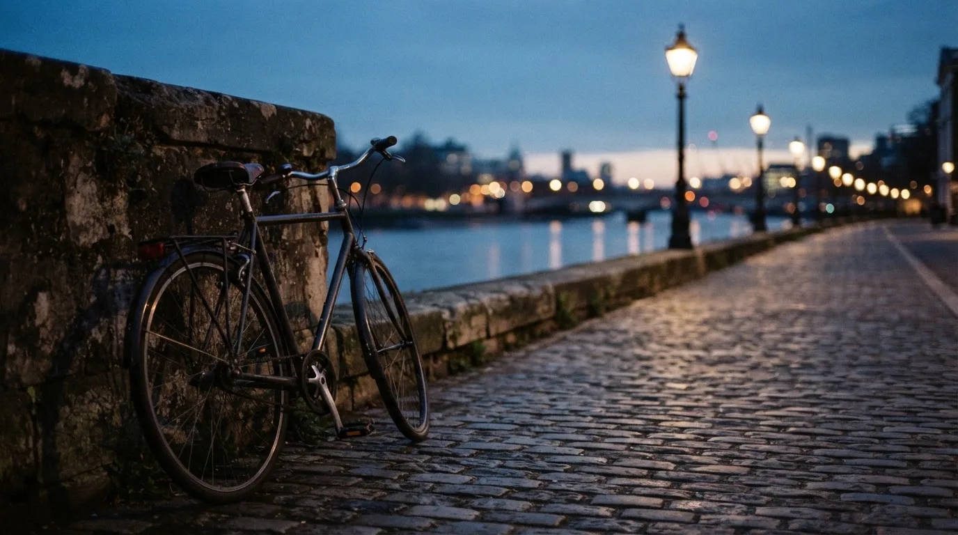Ein Velo lehnt an einer Steinmauer am Ufer eines Flusses, die Strassenlaternen leuchten in der Dämmerung.