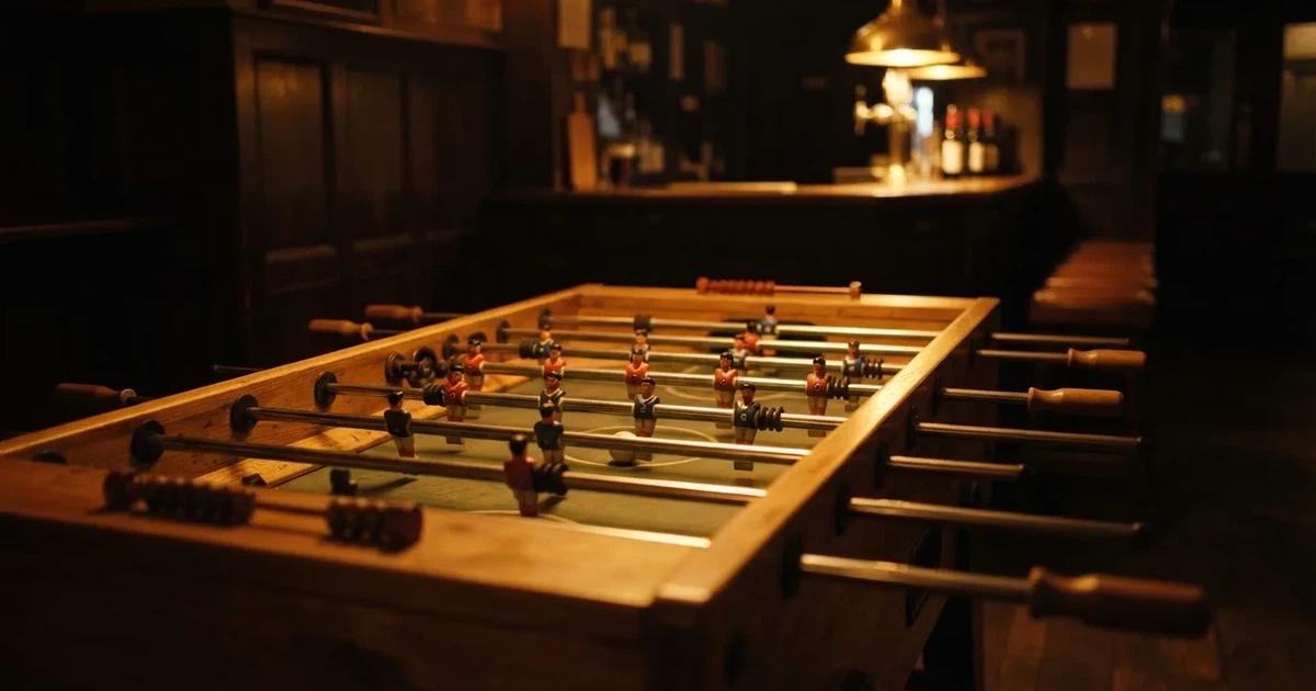 Ein hölzerner Tischfussball steht in einer leeren Bar, die Stangen mit den Figuren glänzen im Gegenlicht einer Lampe.