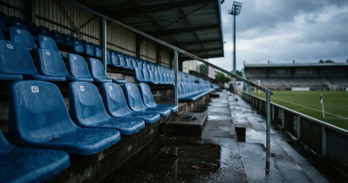 Eine Reihe leerer Sitzschalen auf der Tribüne eines kleinen Stadions, auf den Stufen glänzt Regenwasser.