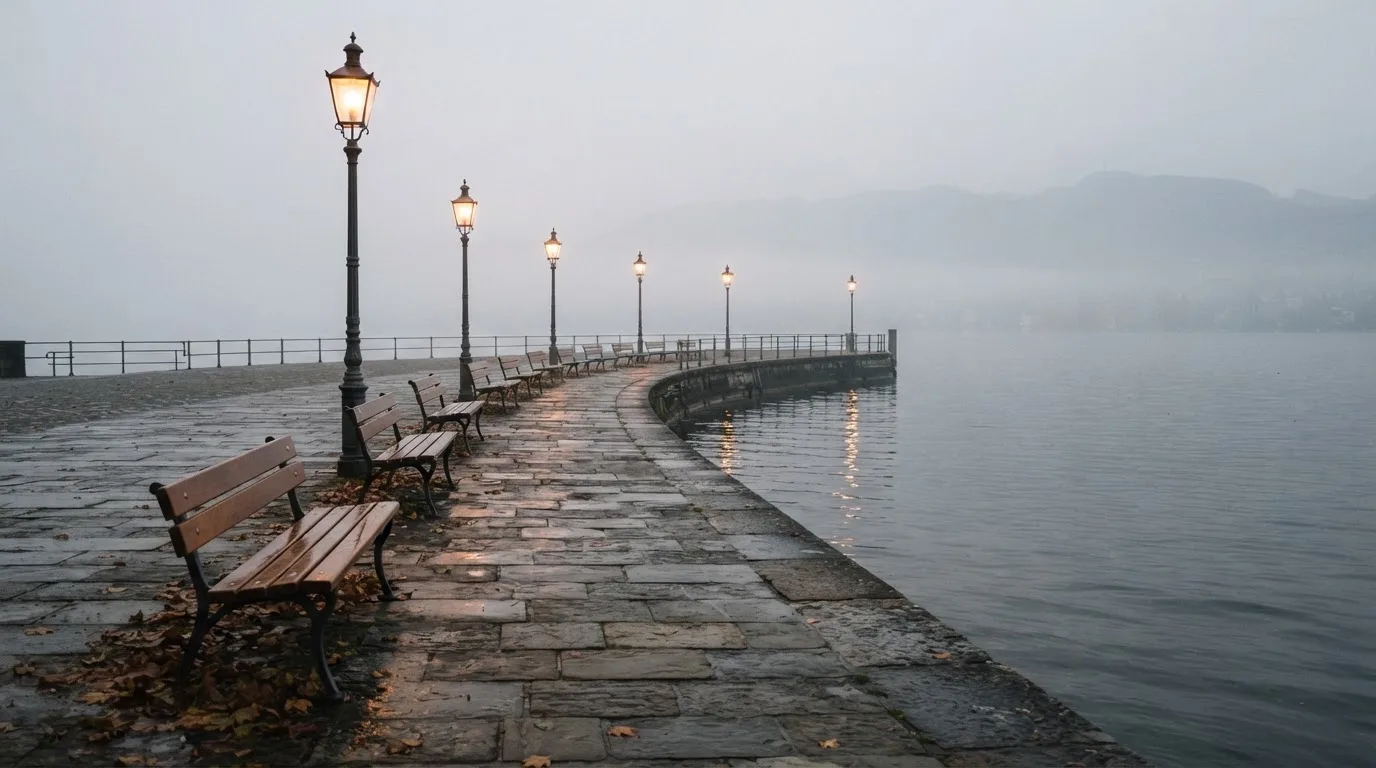 Eine Seepromenade mit leeren Bänken an einem nebligen Morgen, im Wasser spiegeln sich die Lichter der Uferlaternen.