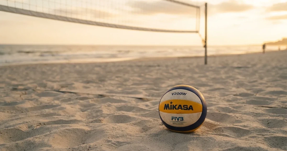 Ein Volleyball liegt auf feinem hellem Sand, ein gespanntes Netz verschwimmt dahinter, das Licht kommt von einem weiten Meer.