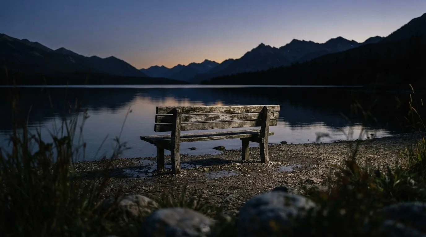 Eine leere Holzbank am Seeufer in der Dämmerung, im Hintergrund dunkle Berge
