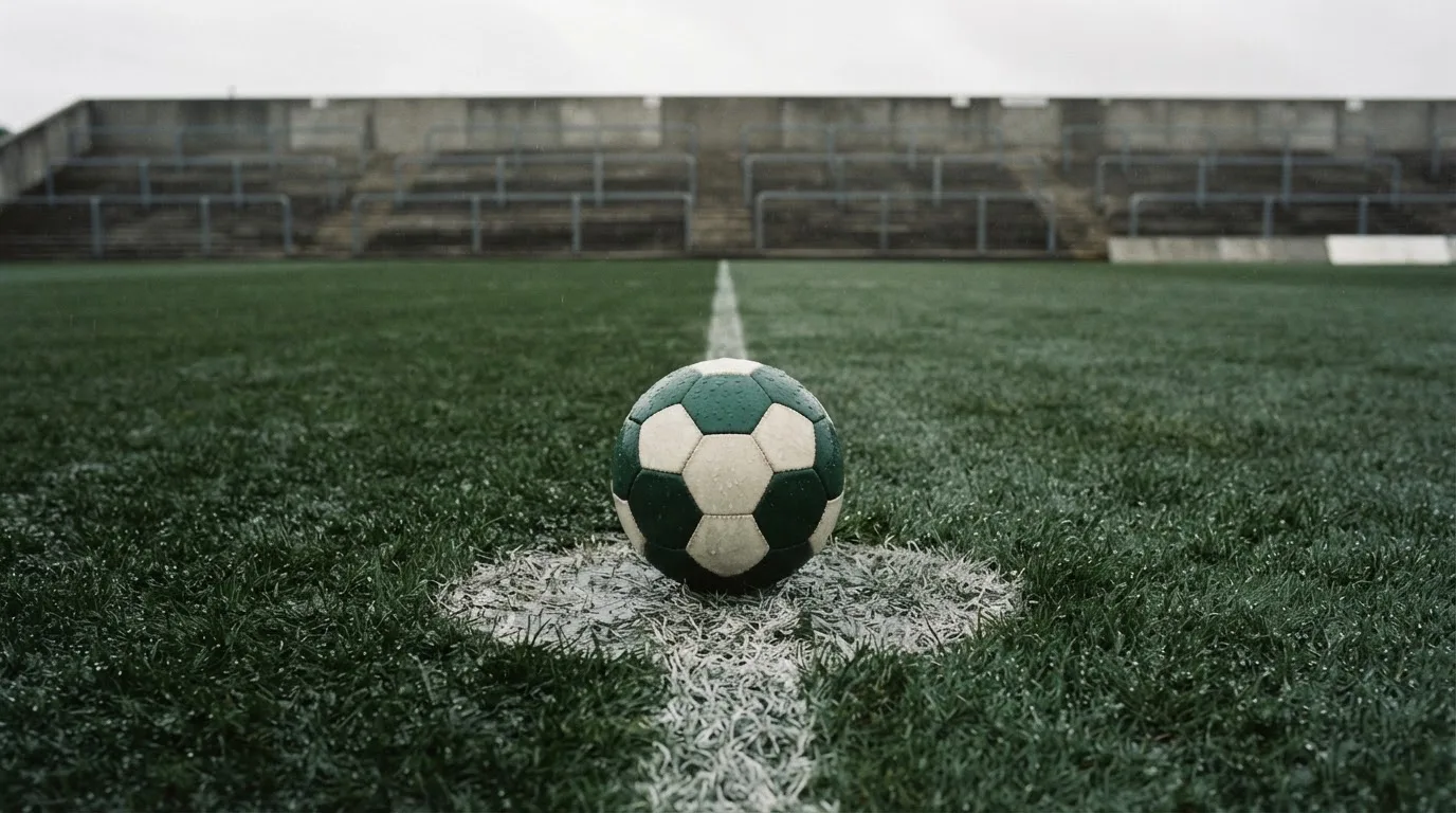 Ein Fussball liegt auf dem Anstosspunkt eines nassen Rasens, dahinter eine leere Tribüne unter grauem Himmel.