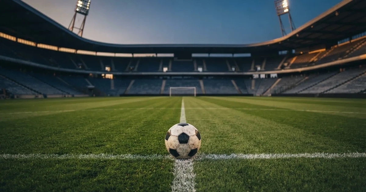 Ein Fussball liegt auf dem Mittelpunkt eines leeren Rasens, im Hintergrund steigen steile Tribünen in der Dämmerung auf.