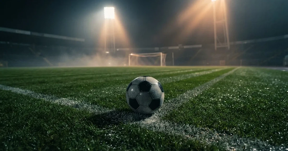 Ein Fussball liegt auf der Strafraumlinie eines nassen Rasens, dahinter stehen Flutlichtmasten im aufsteigenden Nebel.