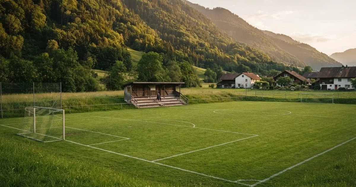 Ein kleiner Fussballplatz mit einfacher Holztribüne und Rasenstreifen am Rand einer Ortschaft, im Hintergrund ein bewaldeter Hügel im Abendlicht.