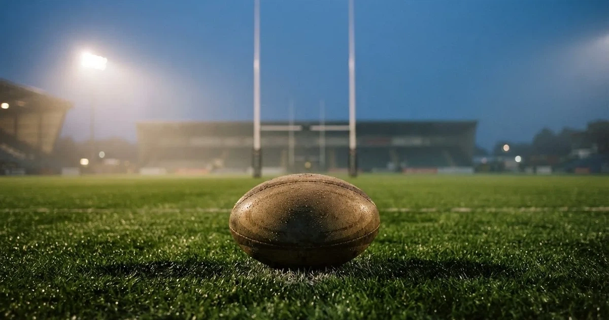 Ein ovaler Rugbyball liegt auf feuchtem Rasen vor zwei hohen Torstangen, hinter dem Spielfeld steigen Nebelschwaden auf.