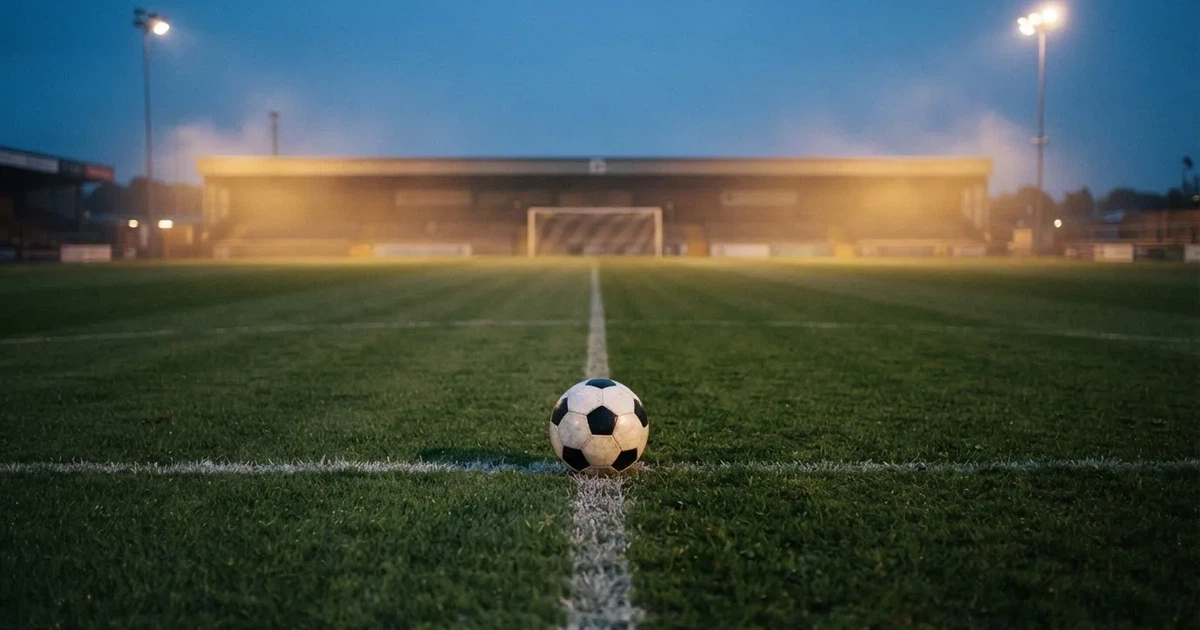 Ein Fussball liegt auf dem Anstosspunkt eines leeren Rasenplatzes in der Abenddämmerung, dahinter leuchten Flutlichtmasten über einer Tribüne.