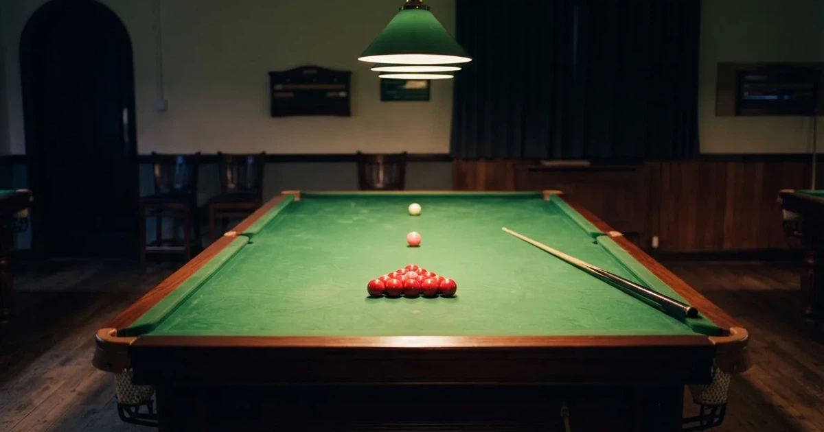 Ein Snookertisch mit grünem Tuch und einem Dreieck aus roten Kugeln unter einer tief hängenden Lampe in einer leeren Halle.