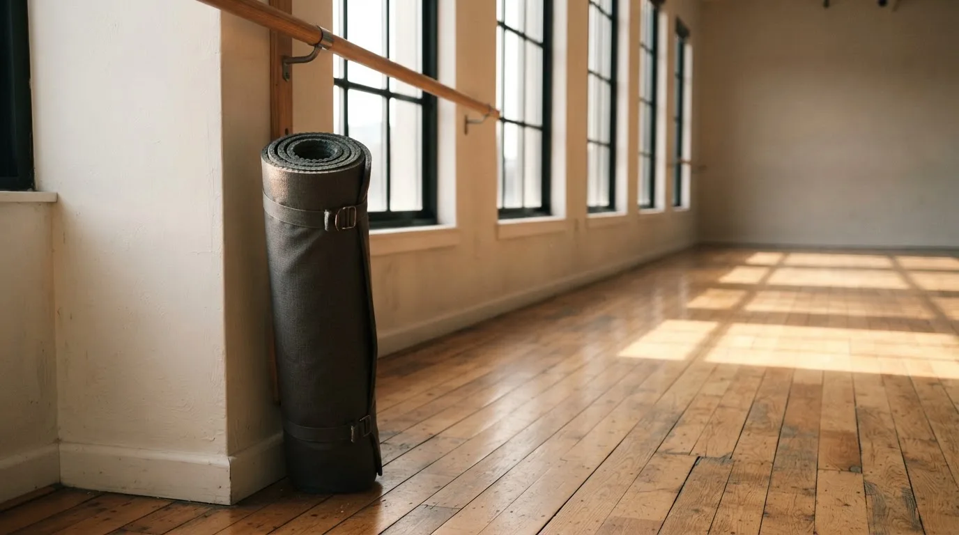 Eine zusammengerollte Trainingsmatte in der Ecke eines leeren Fitnessraums, durch hohe Fenster fällt Morgenlicht.