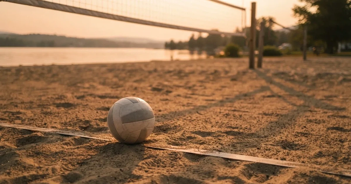 Ein Volleyball liegt im Sand eines Beachplatzes, zwischen zwei Pfosten ist ein Netz gespannt, tiefe Abendsonne wirft lange Schatten.