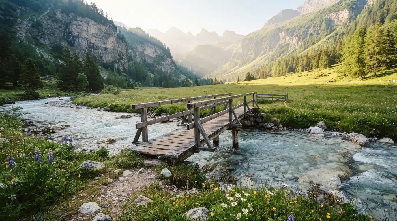 Ein Holzsteg führt über einen klaren Bergbach, im Hintergrund Wiesen und steile Hänge im Morgenlicht.