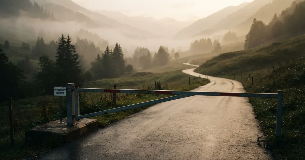 Eine geschlossene Schranke steht an einer ruhigen Bergstrasse im Morgennebel, dahinter Tannen und eine nasse Wiese.