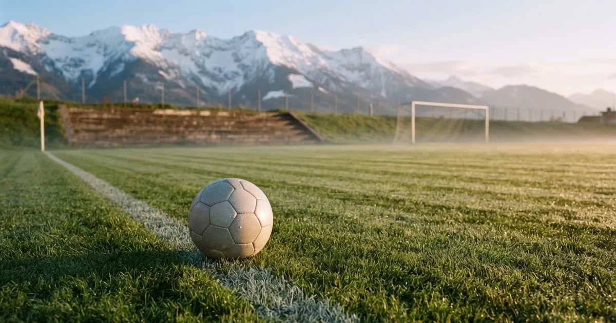 Ein Fussball liegt am frühen Morgen auf dem Rasen eines leeren Fussballfeldes, dahinter eine kleine Tribüne und schneebedeckte Berge.