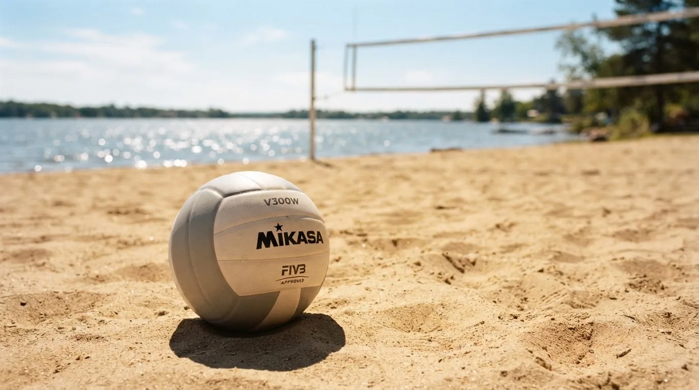 Ein Volleyball liegt auf dem Sand eines Strandfeldes am Seeufer, im Hintergrund glitzert das Wasser.