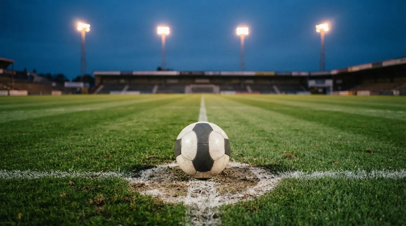 Ein Fussball liegt auf dem Mittelpunkt eines leeren Rasenplatzes, im Hintergrund ragen Flutlichtmasten in die Abenddämmerung