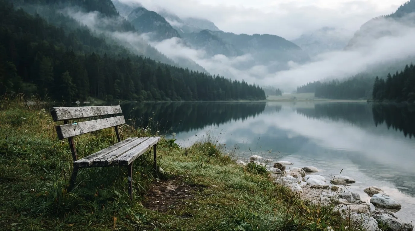 Eine Holzbank am Ufer eines ruhigen Bergsees, am gegenüberliegenden Ufer steigen dunkle Hänge in den Frühnebel