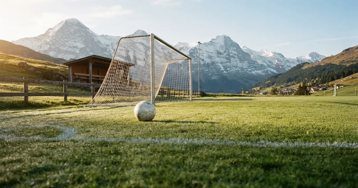 Ein Dorf-Sportplatz mit einem Ball am Strafraumrand, dahinter schneebedeckte Alpengipfel im klaren Morgenlicht