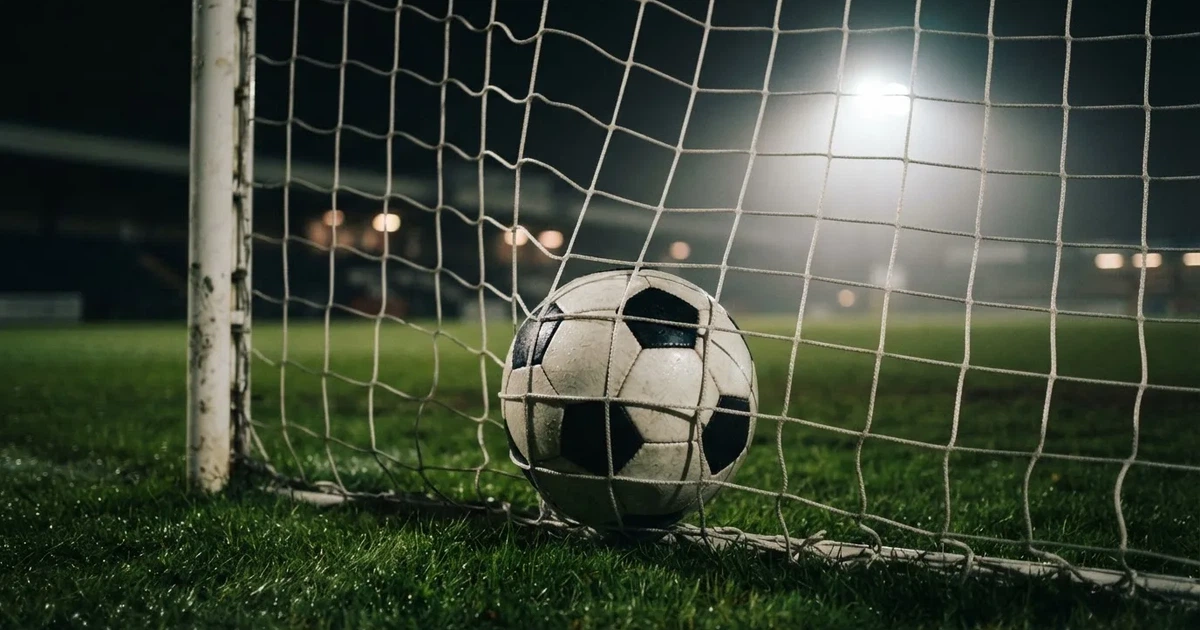Ein Fussball liegt im Netz eines Tores, Flutlicht und kalte Abendluft liegen über dem leeren Platz