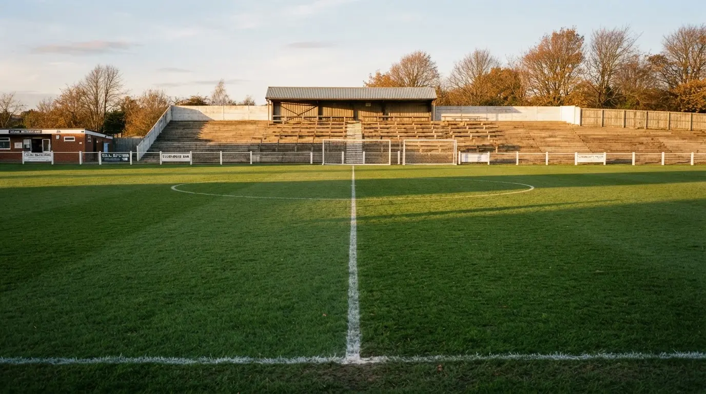 Leere Zuschauerränge eines kleinen Stadions im Herbstlicht, davor ein Rasenplatz mit frisch gezogenen Linien.