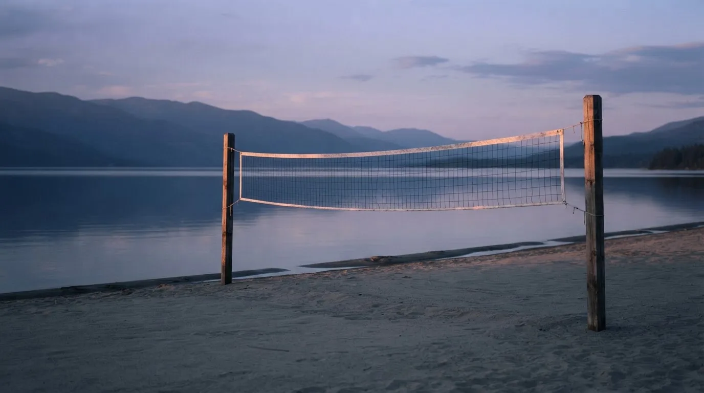 Ein Volleyballnetz steht an einem Seeufer in der Abenddämmerung, der Sand ist leer und das Wasser glatt.