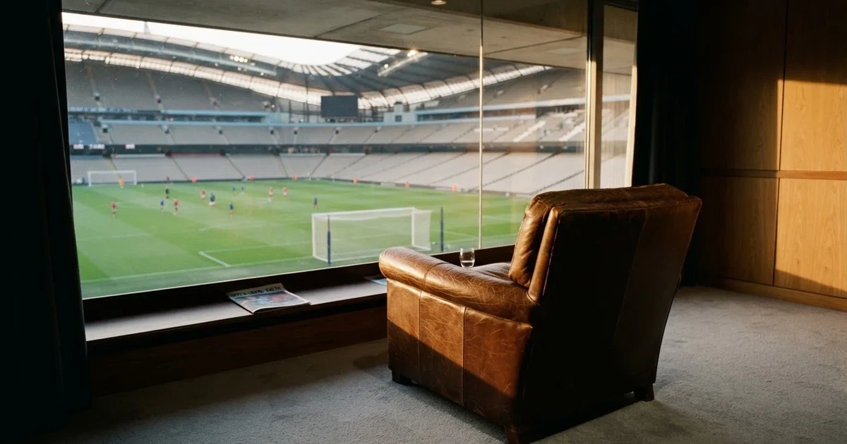 Ein leerer Ledersessel steht in einer Stadionloge mit Blick auf das Spielfeld im Abendlicht.