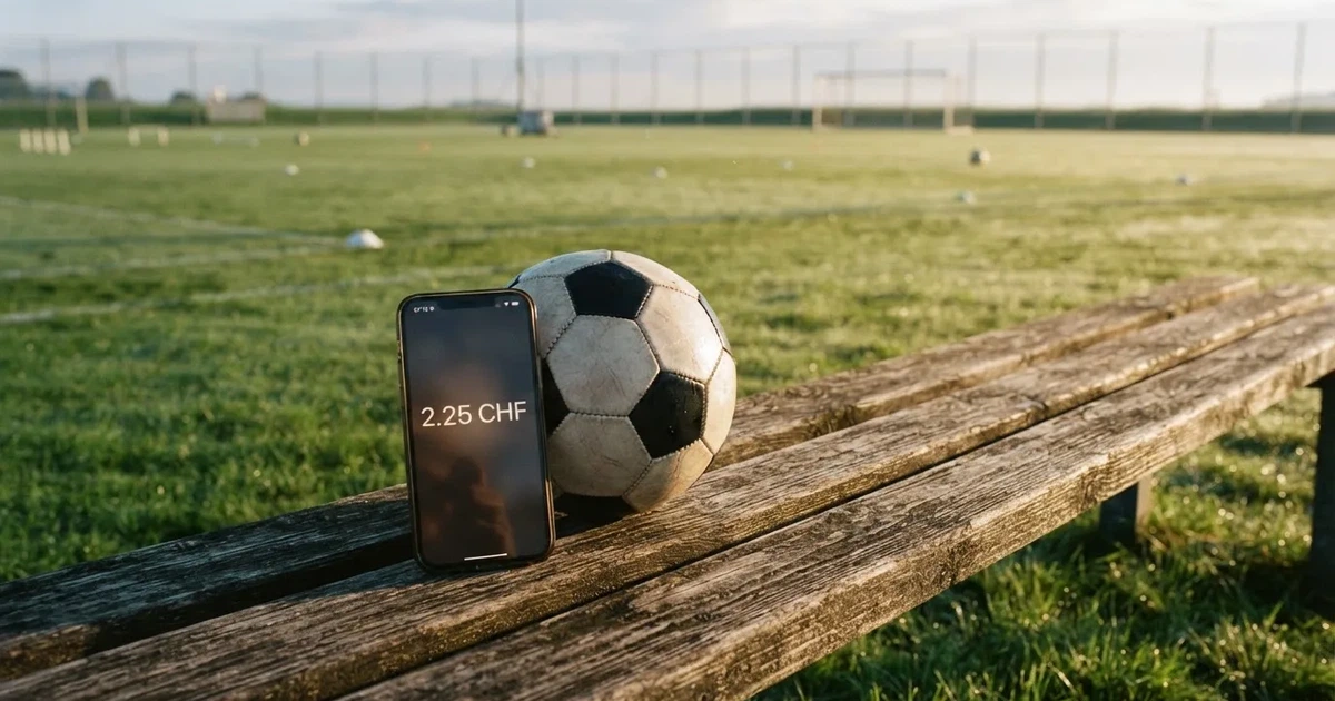 Ein Smartphone mit dunklem Bildschirm liegt neben einem Fussball auf einer Holzbank am Rand eines Trainingsplatzes im Morgenlicht.