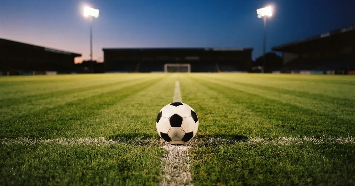 Ein Fussball liegt auf dem Anstosspunkt eines leeren Rasenplatzes, im Hintergrund glimmen Flutlichtmasten im Abendlicht.