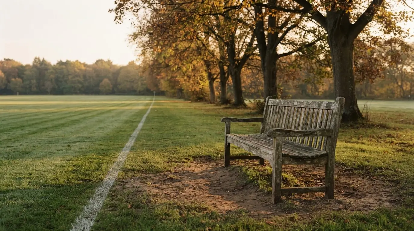 Eine hölzerne Bank steht an der Seitenlinie eines leeren Sportplatzes, dahinter Bäume im Herbstlicht.